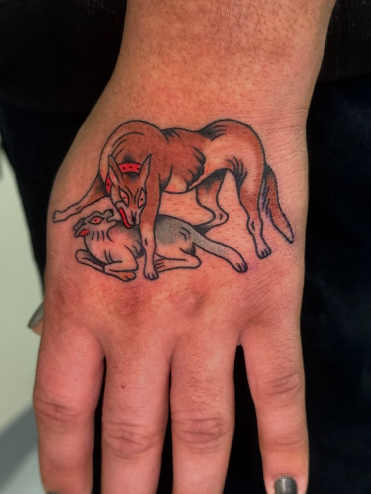 Tattoo on a Latino person’s hand - two dogs, one in a protective stance (brown body, red collar, aggressive expression) over a smaller dog (gray body, looking surprised) tattoo is in color and fills most of the top of the hand 