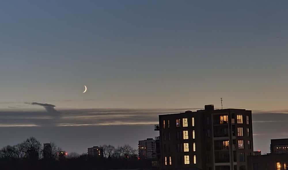 A photo of the sky with a sliver of moon. Also a building, most of its windows lit up, and bare leafless trees.