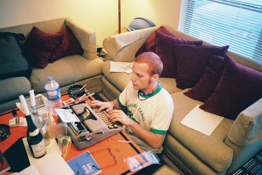 A young man in his 20s sits at a typewriter. He has short, very short hair, dyed red. Wears a green "ringer" t-shirt that says "Panama Canal." He has long sideburns, but they are trimmed close to his face. He sits on the floor, leaning against a loveseat, which is at a right angle to an identical loveseat. Both loveseats are covered with maroon pillows, though on the loveseat to his right is also a dark article of some sort — perhaps a jacket, a blanket, or another pillow. In the corner, in the space where the two loveseats meet, there is a floor lamp (one can only see, however, the lamp's stand-pole, and the evidence of its light on the wall behind it). In the same corner one can see the top of a rubber trashcan, like one might find in an industrial shop (not typical for a home). The typewriter is on a coffee table that is made of a stolen street sign or construction sign, orange, maybe "Yield," maybe "Road Closed." A cigarette burns in an ashtray to his right. Scattered around him are sheets of notebook paper with ink drawings on them. On the table are also two wine bottles with candles protruding, a closed notebook of sketch paper, a slip of plastic, a few empty-looking soft-packs of cigarettes, various pens and pencils and a paintbrush, and a pair of rose-colored sunglasses, lying upside down, hinges fully open, temple tips facing upright. Behind the loveseat is a set of blinds, behind the set of blinds is a window, through which is visible daylight and the front of a car. It is not possible to read the car's license plate. In the typewriter, the paper the young man types onto is a lined sheet from a spiral pad. The words he types are framed, hemmed in at the top by ink drawings, black looping lines inside rounded rectangles, and flanked to the left by the fluttering tattered edges where the paper was torn from the corkscrewing metal that once served as its notebook's spine.