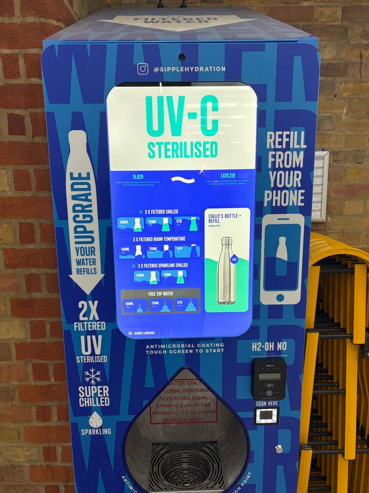 A bright vending machine reads “UV-C Sterilised” “Refill from your phone” “Upgrade” “2x filtered” “H2-OH NO” above a credit card reader device). A touch screen displays a plethora of different options for water refills. Brightly coloured options propose 3x filtered chilled 500ml - 45p, 750ml - 60p, 1000ml - 75p, At the bottom in dull grey “free tap water” 500ml, 750ml, 1000ml