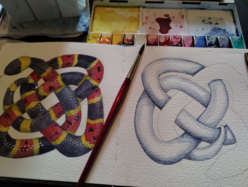 Open sketchbook with watercolor tools by it. The left page has a completed painting of an Eastern Coral Snake twisted into a Celtic knot. The right page has the outline of a different knot shaped snake which has been partially shaded in indanthrone blue.