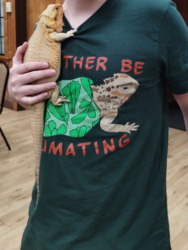 Torso of a white dude holding a very round and docile bearded dragon. He is wearing a forest green t-shirt with an illustration of a greater short horned lizard wearing a leafy blanket and the partially obscured words "I'd rather be brumating".