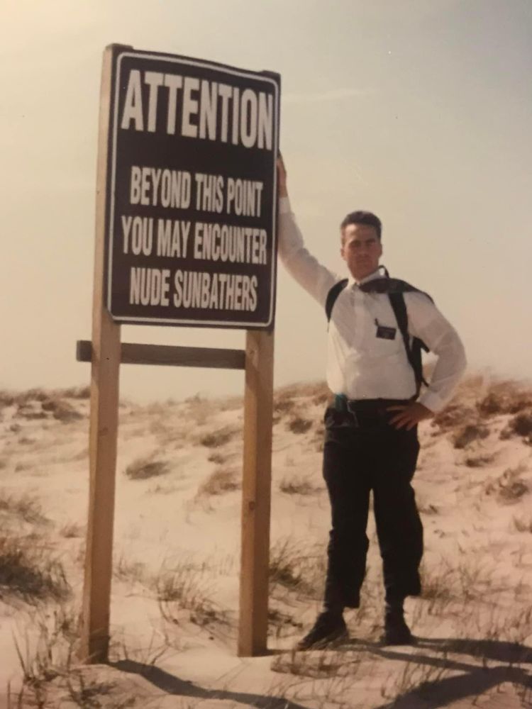 A Mormon missionary stands in a windy beach next to a sign which reads “attention: beyond this point you may encounter nude sunbathers”