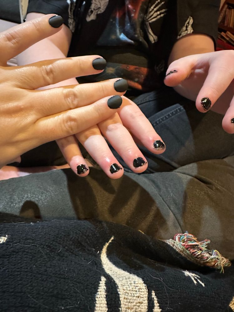 An adult hand with matte black nails is resting palm down on top of a child’s hand with glossy black nails. The other child’s hand is held off to the side. A black and white quilt is in the background on the bottom of the photo. 