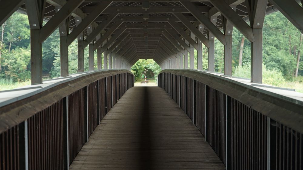 sehr symmetrisches Foto einer Holzbrücke für Fußgänger (und eventuell Radfahrer)
die Holzbrücke ist überdacht und auf dem Foto sieht man sowohl die Holzelemente der Seitengeländer als auch die die Sparren der Dachkonstruktion
in der Mitte ein dunklerer Holzstreifen am Boden der genau auf das Bildzentrum in Form eines rot-weissen Pfahles zuführt. Links davon ein grüner Abfalleimer