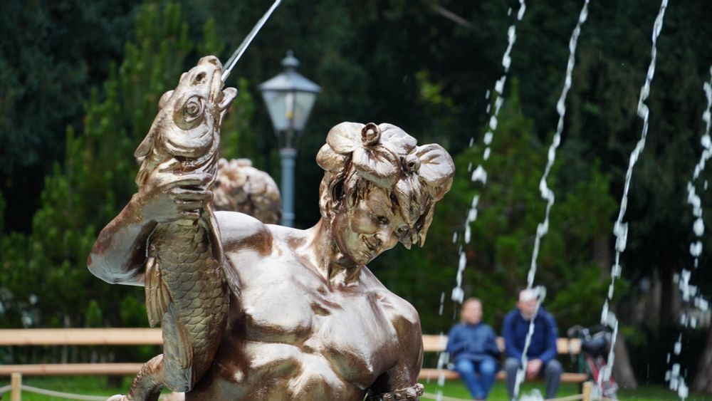 eine frisch renovierte, metallisch glänzende Brunnenfigur mit nacktem Oberkörper hält einen wasserspeienden Fisch in der Hand
Im Hintergrund vor grün/dunkelgrünen Bäumen sieht man eine Laterne und zwei blau gekleidete Menschen auf einer Bank sitzend
4 schräge Wasserstrahlen wie eingefroren im rechten Drittel des Bildes