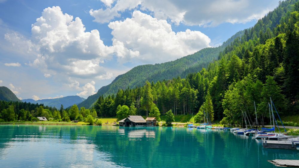 Uferlandschaft
bewaldete Höhenrücken streben vom Bildzentrum links nach rechts oben
Wolken und Wald spiegeln sich im Wasser
Hütten und Sehelboote am Ufer
