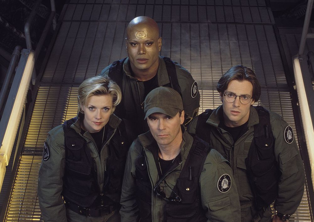 The crew of the sci-fi television series "Stargate SG1" at the foot of the ramp that leads up to the titular Stargate. They include, going clockwise from top, Teal'c, Daniel Jackson, Jack O'Neill, and Samantha Carter. 