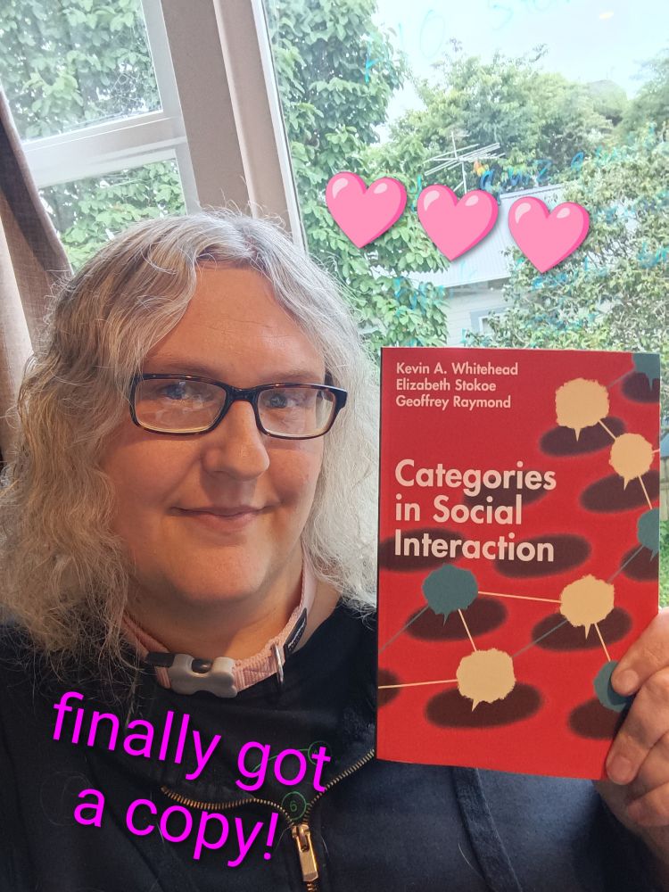 Picture of me holding up a copy of a book "Categories in Social Interaction" up in front of me. Above the book are three pink emoji hearts. Below is the text "finally got a copy!" Behind me is the garden outside my window.