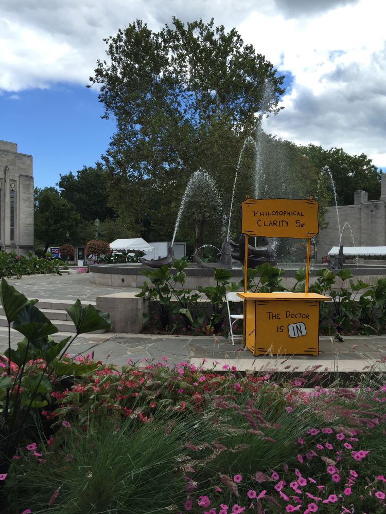 Springtime photo of Showalter Fountain at Indiana University Bloomington. A bright yellow stand advertising "philosophical clarity" for 5 cents is in the foreground with the words "The doctor is in" below, but the seat behind the stand is empty.