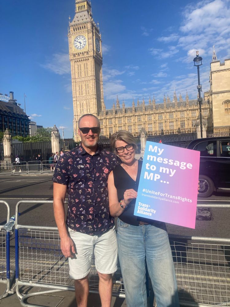 Me and my husband standing in front of Big Ben and the Houses of Parliament. It’s sunny and I’ve forgotten my sunglasses so i’m squinting into the sun. I’m carrying a placard in trans colours “My message to my MP…” 