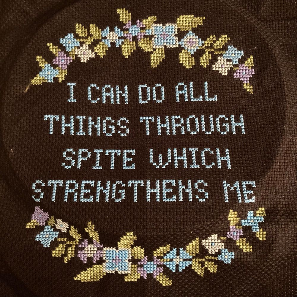 Crossstitch of blue text on black aida reading “I can do all things through spite which strengthens me”