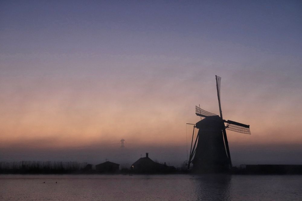Optrekkende mist, een grijze band tegen de horizon en uitwaaierend, tegen een blauw en roze zonsopgang. Silhouet van molen en bijgebouw op links van de molen.