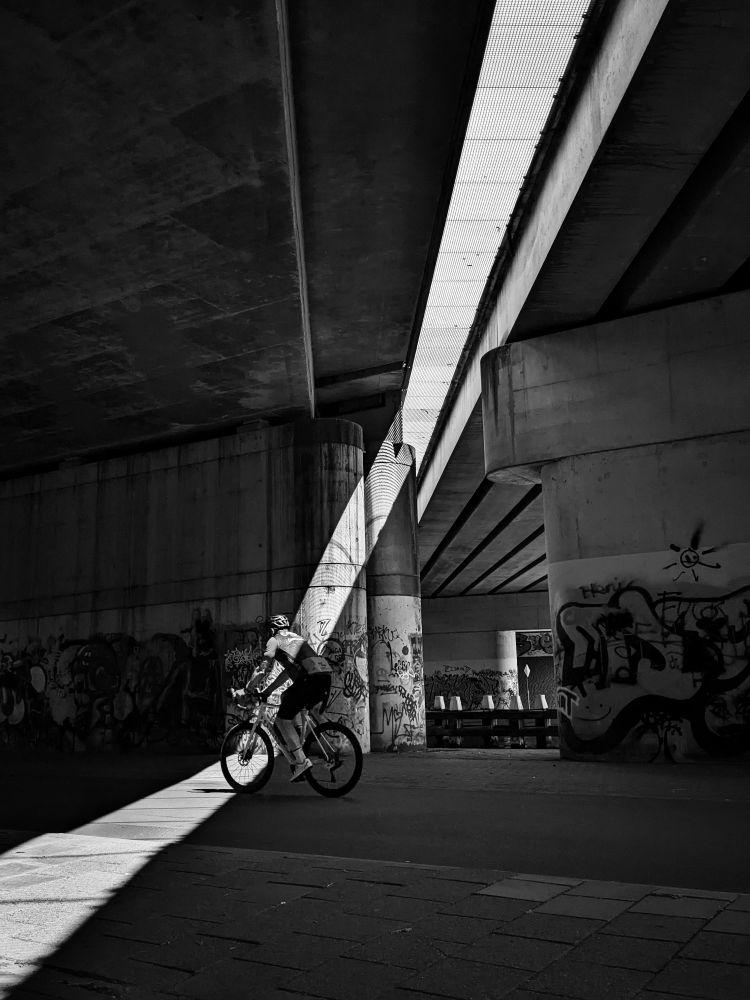 A streak of sunlight coming through an opening in a bridge, shines on a bike path beneath it. A cyclist just passing through it. Behind him some pillars with graffiti and white capped bollards. Openings, looking like stripes,  between the concrete beams at the underside of the bridge