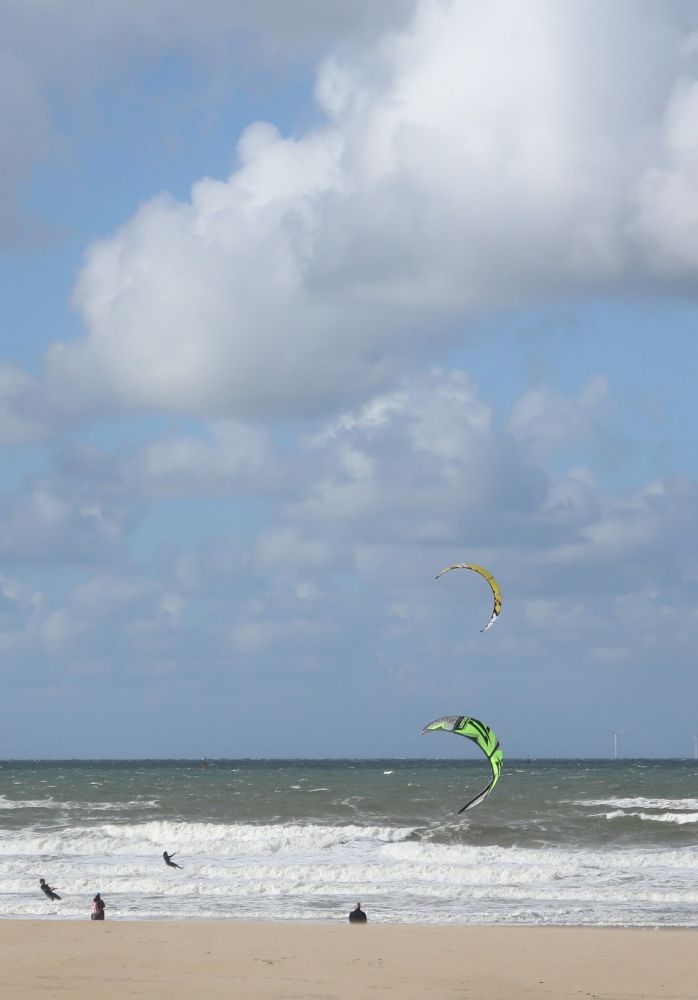 Two people on the left kite surfing in the surf of a blue, green looking North Sea. Blue sky with big fluffy white clouds. Bright green and yellow kites. Some spectators on the sandy beach.