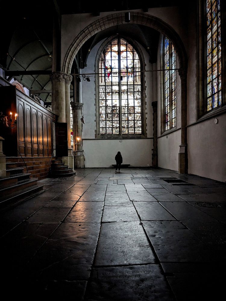 Sint-Janskerk in Gouda. Light through stained glass windows casts a soft glow on a granite floor. Small artificial candles throw a soft golden glow on the wood work on the left. A person standing in front of the window at the far end beneath an arche.