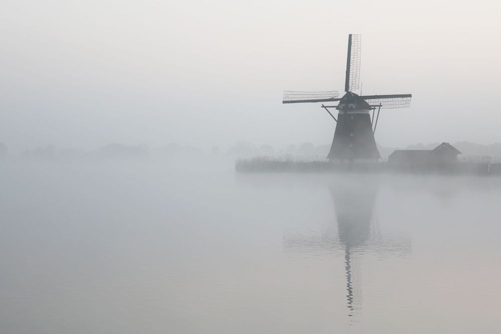 Molen op dijk op rechts in de mist reflectie in het water. De rest van de foto een witte wereld.