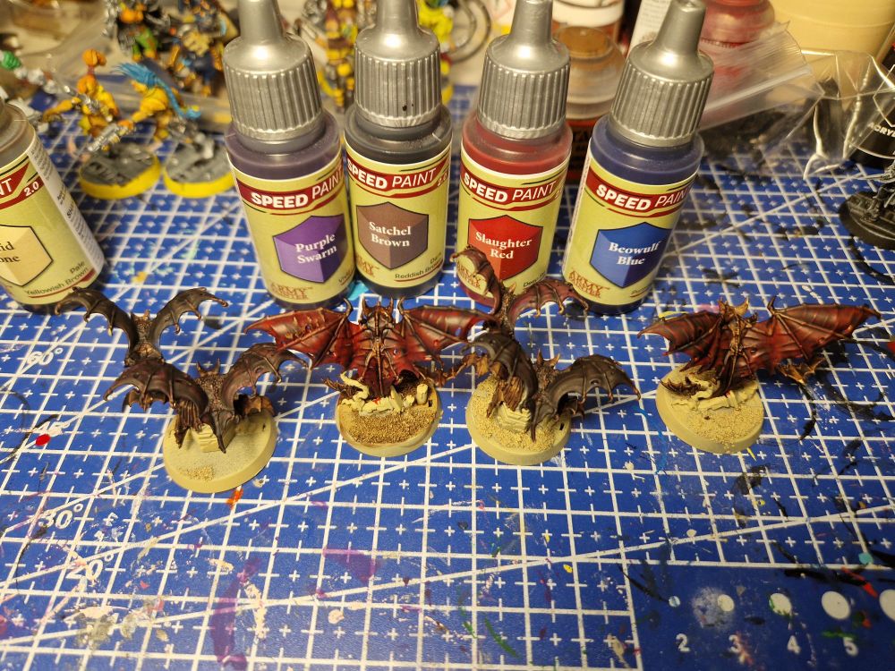 Line up of 4 model bats, from an overhead viewpoint. The first and third models have 2 medium sized bats and the second and fourth have one large bat. All 4 have smaller bats hanging around but are not all visible. Also in the picture are 4 paints - from the Army Painter's speed paint range. We have purple swarm, satchel brown, slaughter red and beowolf blue. The two sets of medium bats are painted in either purple or blue, with a brown coar over the top. The big bats are painted in red with a brown coat over the top. Any visible small bats are just painted in brown.