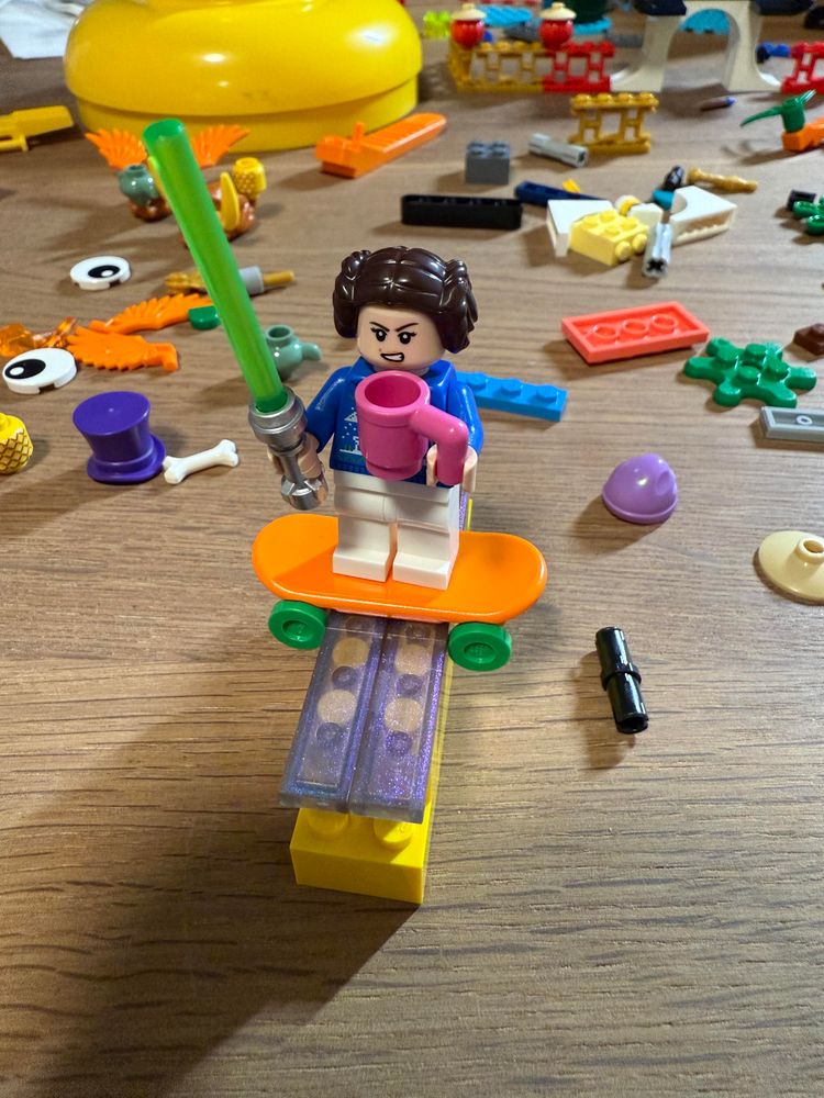 A Leia minifigure holding a pink mug and a green lightsaber, on an orange skateboard with green wheels, sitting atop a yellow wall with translucent covering. Lots of random Lego pieces are scattered around. 