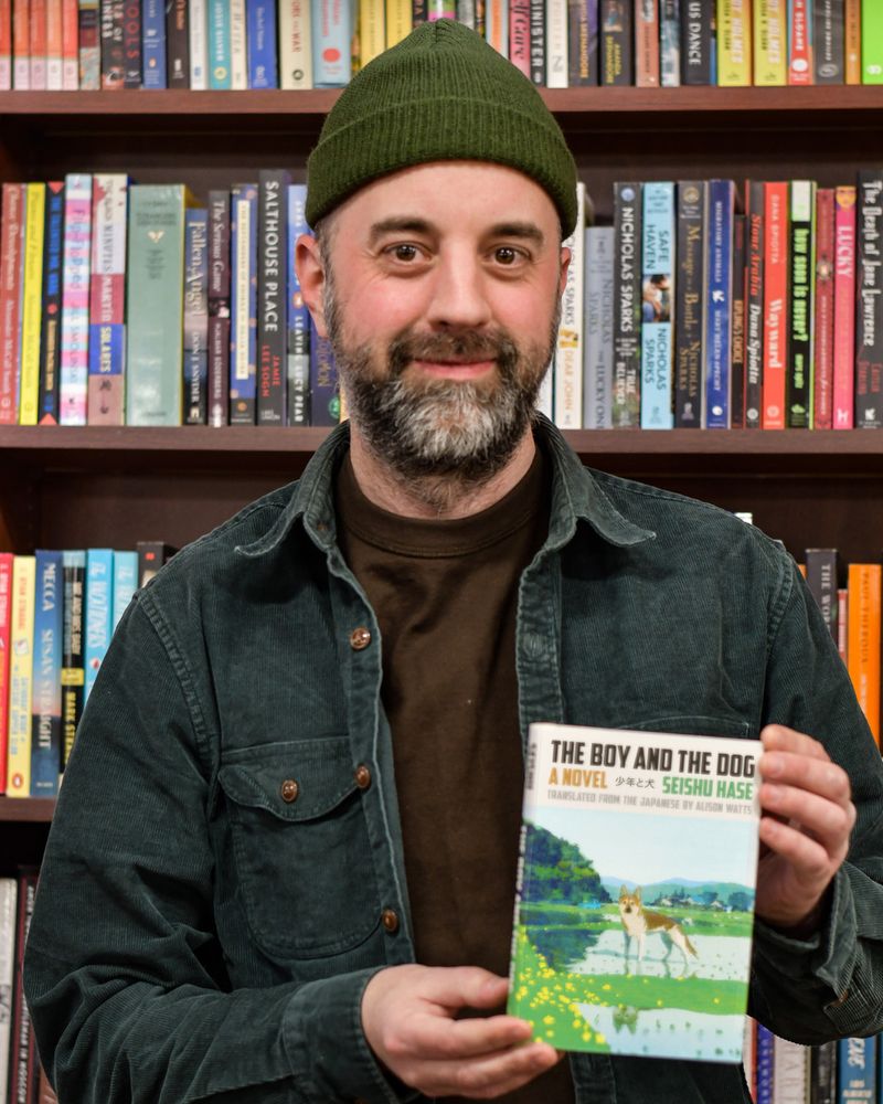Image of a man in a green beanie standing in front of bookcases and holding a copy of THE BOY AND HIS DOG by Seishu Hase; book cover shows a dog facing the reader, standing in a green and watery landscape.