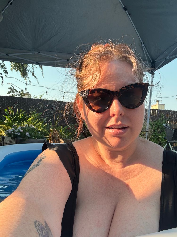 Red haired woman wearing sunglasses with the hint of a blowup pool peeking behind her. Sun shining down, casting shadows across chest, face, and arm.