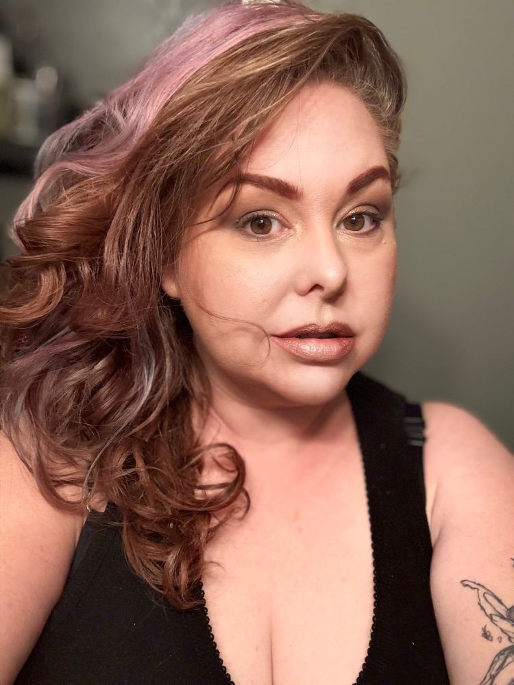 Styles curls in copper and faded shades of violet frame one side of a brown eyed woman's soft face with slightly parted lips.