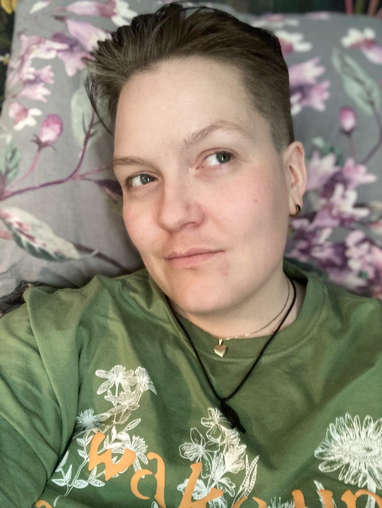 Bilde av en ung mann som ser opp til høyre med et lurt smil på leppene. Hodet er tiltet litt mot venstre. Jeg har på en grønn t-skjorte med hvite tegninger av blomster og oransje bokstaver trykket over det igjen. Håret er barbert kort på sidene og ligger bakover på toppen, med litt volum i front. 