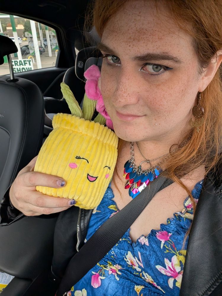 Alannah, in the car while traveling to go see an amazing cute girl, is holding a plushie of a smiling flower pot with pink flowers. She is wearing a blue floral dress and a leather jacket. She has also done her eye makeup.
