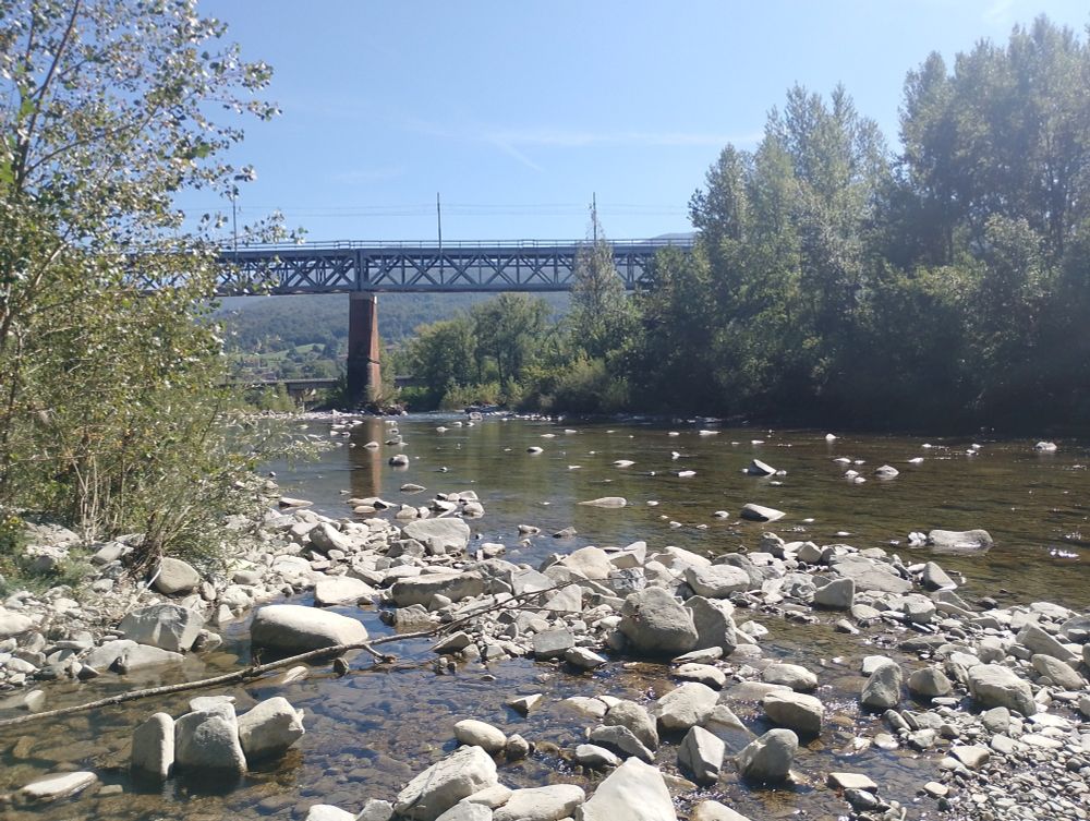 Fiume che scorre, con sassi grigi e bianchi un po' ovunque. In lontananza, si vede il ponte di ferro di una ferrovia.