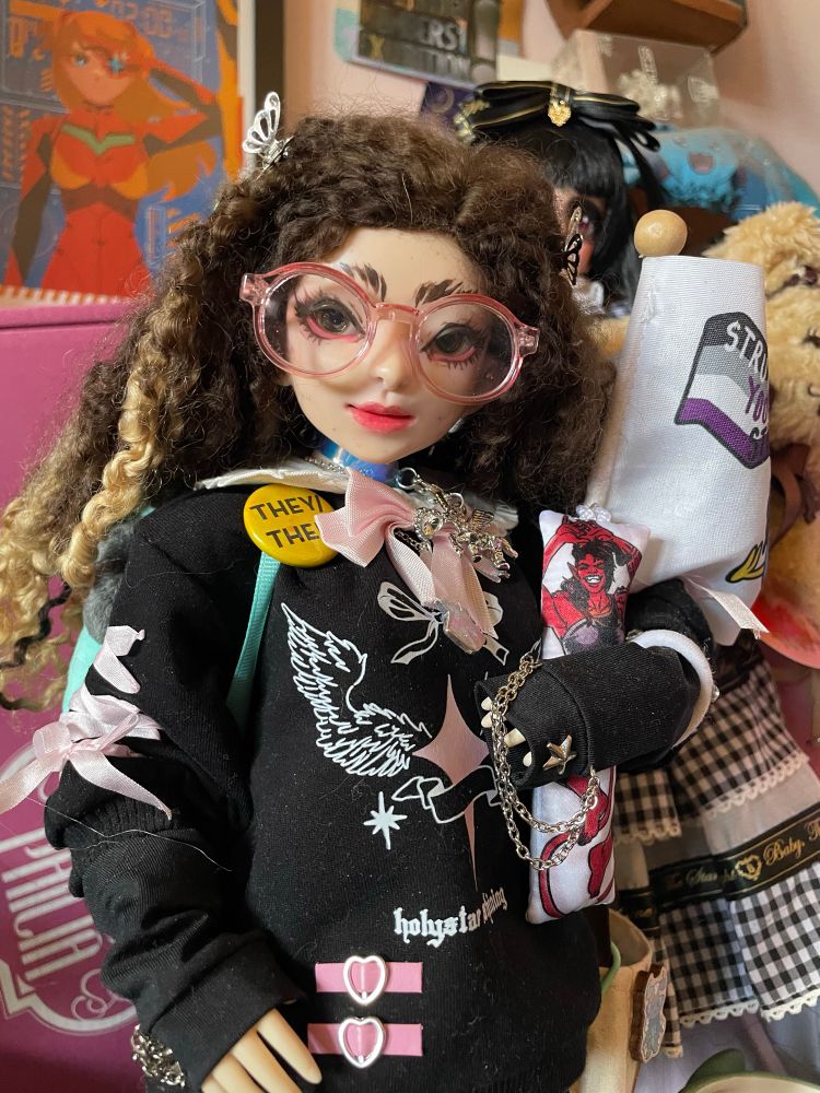 A resin & vinyl hybrid doll. A faewood head on a obitsu body. They are wearing a Jirai Kei styled sweat shirt dress, a they/them pronoun pin and carrying a miniature Karlach body pillow. They have long brown curly hair and wear pink translucent glasses.