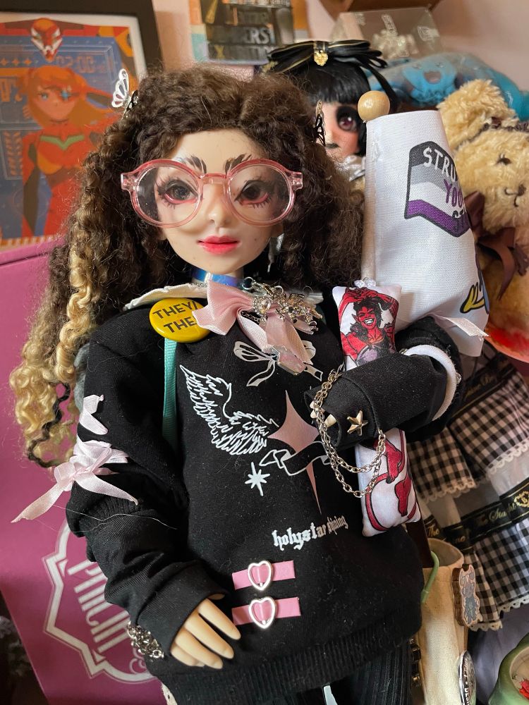 A resin & vinyl hybrid doll. A faewood head on a obitsu body. They are wearing a Jirai Kei styled sweat shirt dress, a they/them pronoun pin and carrying a miniature Karlach body pillow. They have long brown curly hair and wear pink translucent glasses.