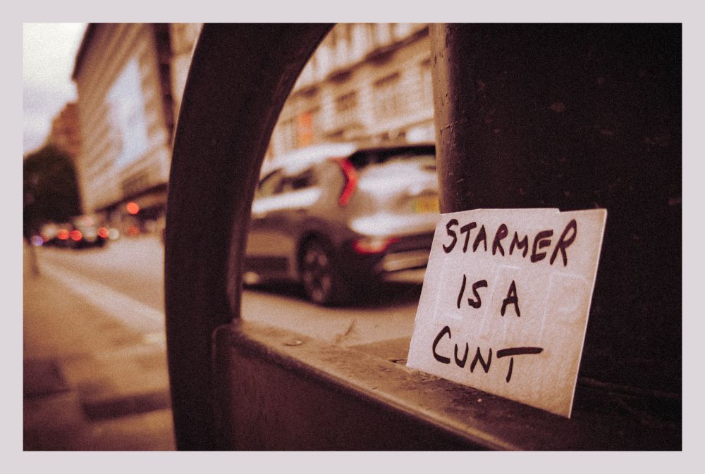 a close-up of a small hand-written note that says "STARMER IS A CUNT" sat on a small bike railing against a background of traffic. the photo is bordered in white and is in a slightly desaturated colour profile