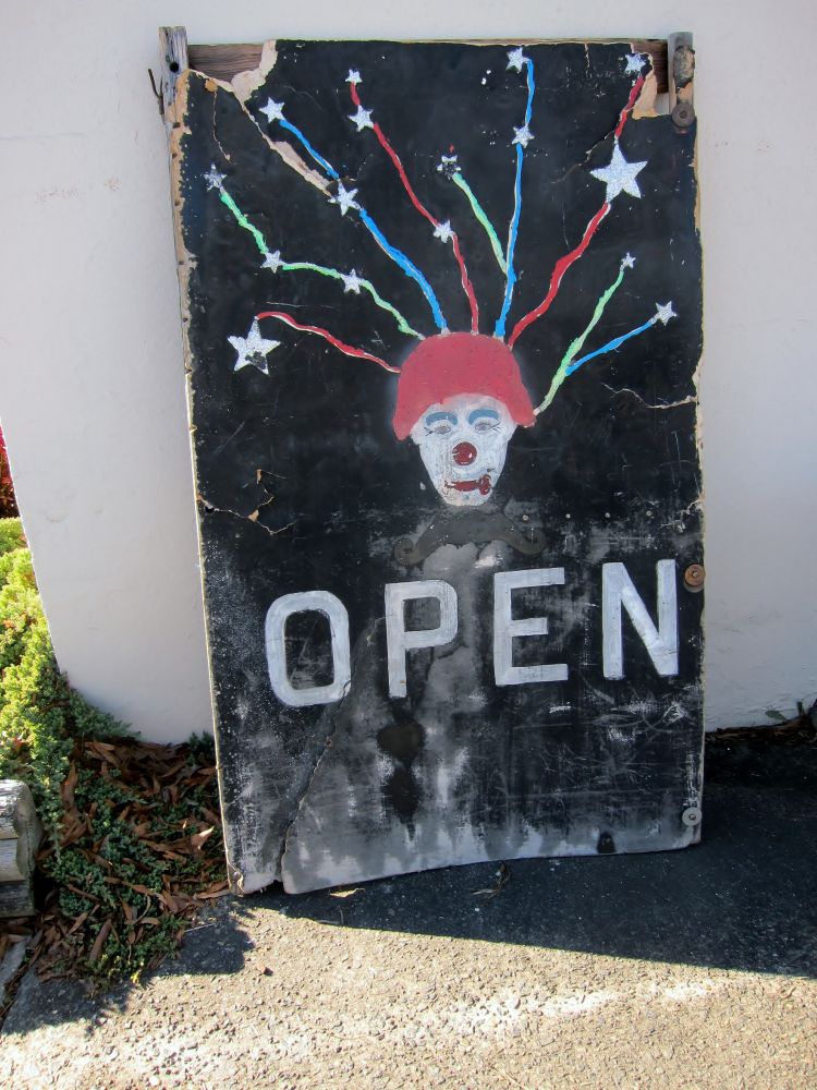 A primitive clown face with a red hat and stars with colorful trails coming out of its head. On a black background. Sign is sitting on the sidewalk outside a business. 