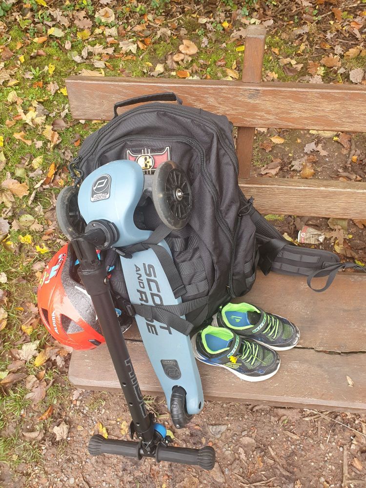 Mein Rucksack steht auf einer Holzbank. An dem Rucksack ist der Roller und Helm des Zwergs angebracht. Davor stehen seine Schuhe.
