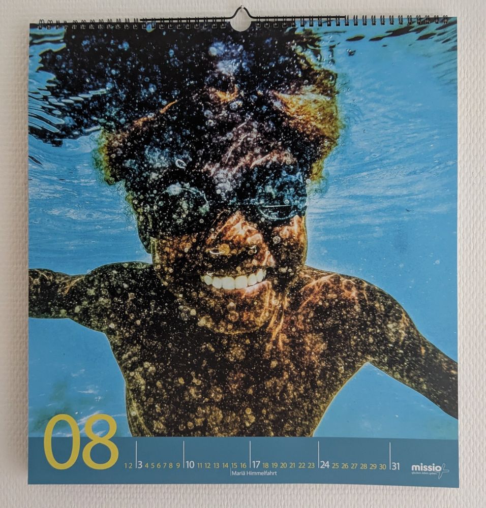 Unterwasseraufnahme eines lachenden Jungen mit dunkler Schwimmbrille, der frontal in die Kamera blickt. Er schwimmt unter der Wasseroberfläche in klarem, blauem Wasser, durch das Sonnenlicht und Luftblasen seinen Oberkörper in ein lebhaftes Muster tauchen. Der Kalender zeigt den Monat August, hervorgehoben mit „08“ und einer Datumsleiste am unteren Bildrand. Am 15. August ist „Maria Himmelfahrt“ markiert. Rechts unten steht das Logo von „missio“. Die Szene wirkt lebendig, fröhlich und sommerlich.