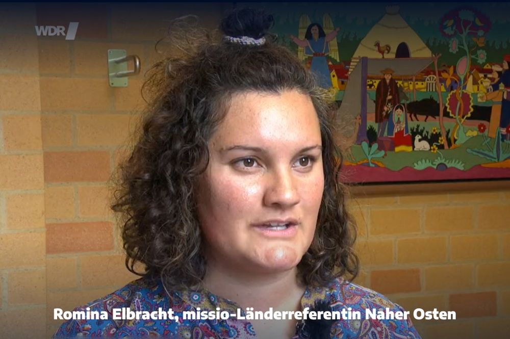 Eine Frau mit lockigen braunen Haaren und buntem Oberteil sitzt vor einer beigefarbenen Backsteinwand. Im Hintergrund hängt ein farbenfrohes Bild mit einem Dorf, Tieren, Pflanzen und Menschen. Unten steht der Text: "Romina Elbracht, missio-Länderreferentin Naher Osten". Oben links das WDR-Logo.