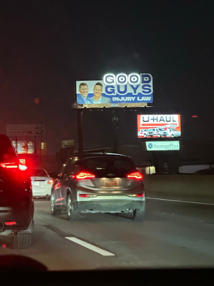 A billboard for 'Good Guys Injury Law' at night. "Good" is composed of lit letters, but the 'd' is burned out, making it look like id reads "Goo Guys"