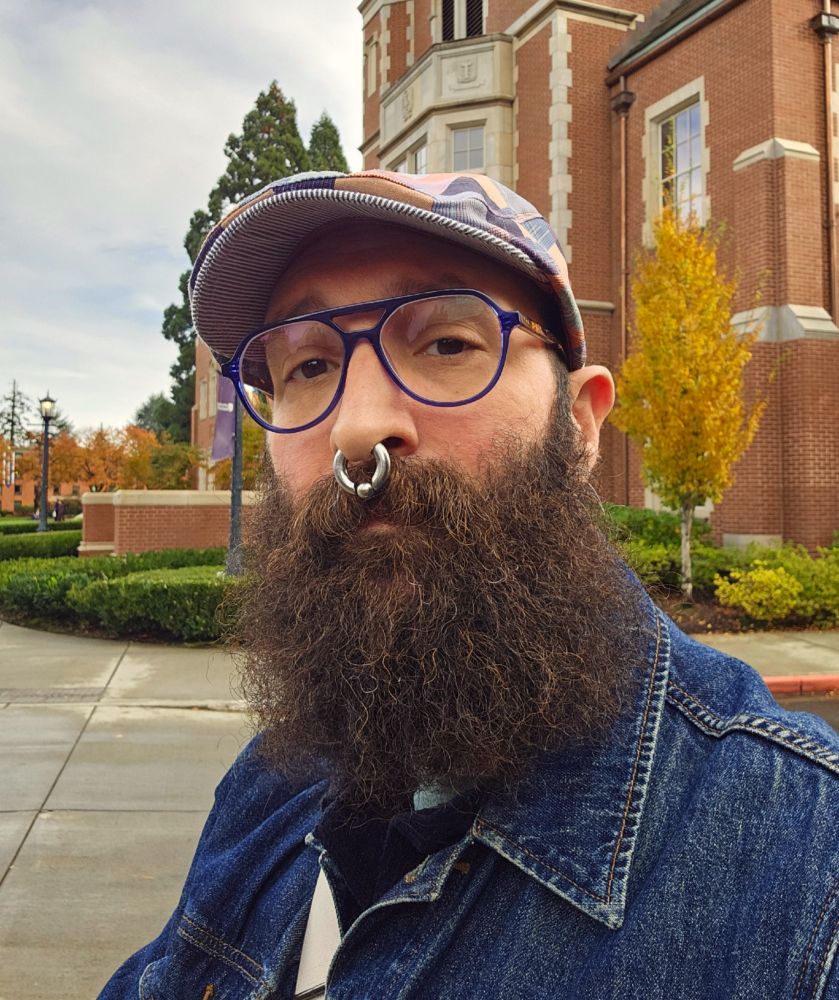 A selfie of me outside with a yellow tree and a brick, quoined university building behind me. I am wearing a denim jacket, blue aviator glasses, and a patchwork cap. 