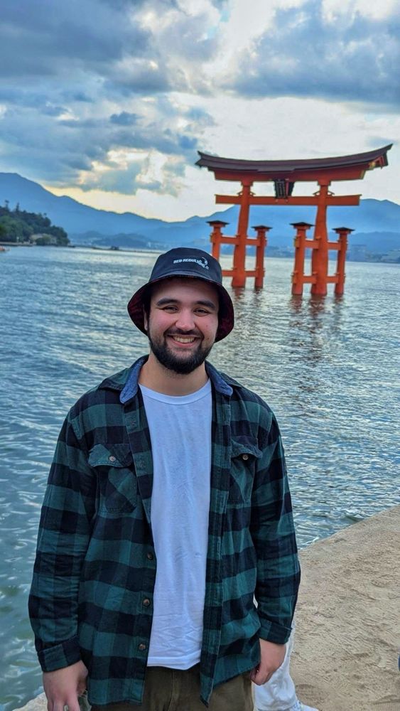 A man in green flannel, a white tee, brown corduroy pants, and a bucket hat smiles at a point overlooking a famous Torii gate in Miyajima, Japan. The gate is partially submerged in water and mountains can be seen in the distance. It's partially cloudy and the sun is beginning to set. This image was modified using an eraser tool to remove two other people who were standing to my left.