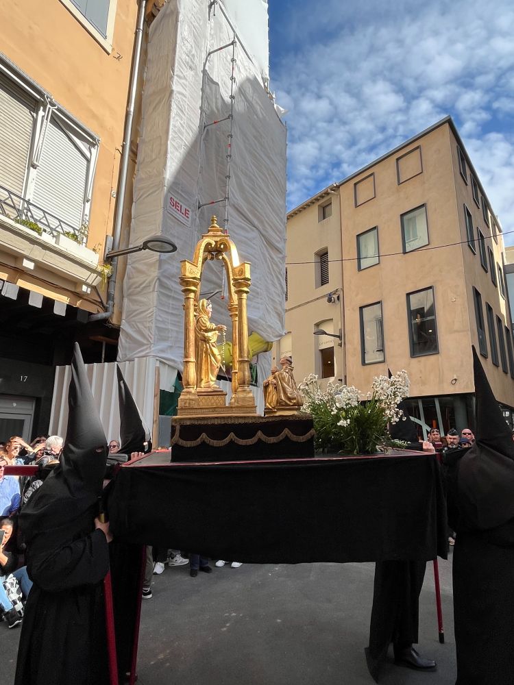 A shot of four black-robed penitents carrying a golden statue of Mary decked with flowers. Somebody, also golden, is kneeling praying to Mary. There are buildings behind them and a bright blue sky with fluffy white clouds up above.