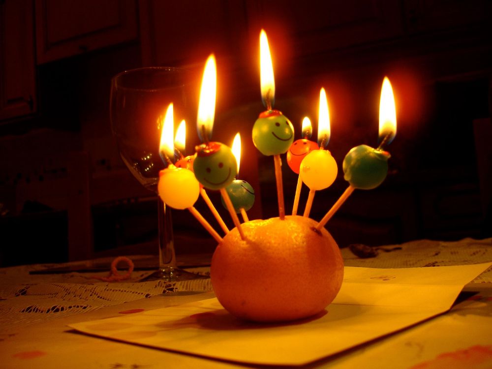 An orange with eight lit birthday candles stuck on it. The candles are yellow, green and red and have happy faces on them. 