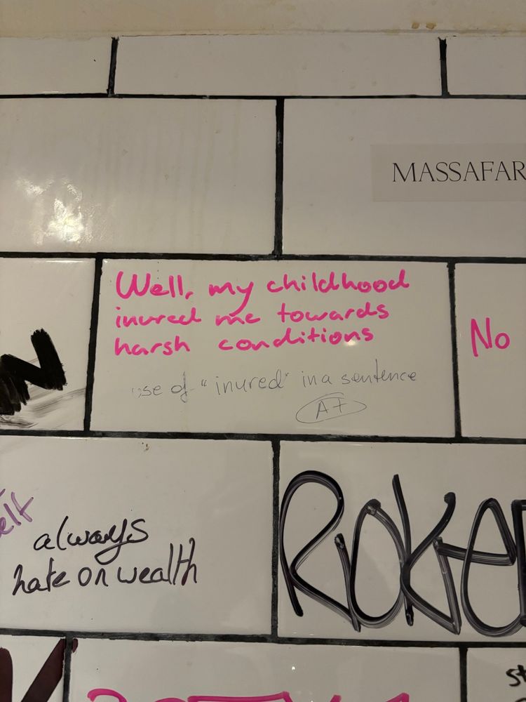 photo of a tile wall with graffiti. the main piece is a sentence written in pink marker that says: “Well, my childhood inured me towards harsh conditions” and beneath it, in different grey writing, is written “use of “inured” in a sentence (A+)”