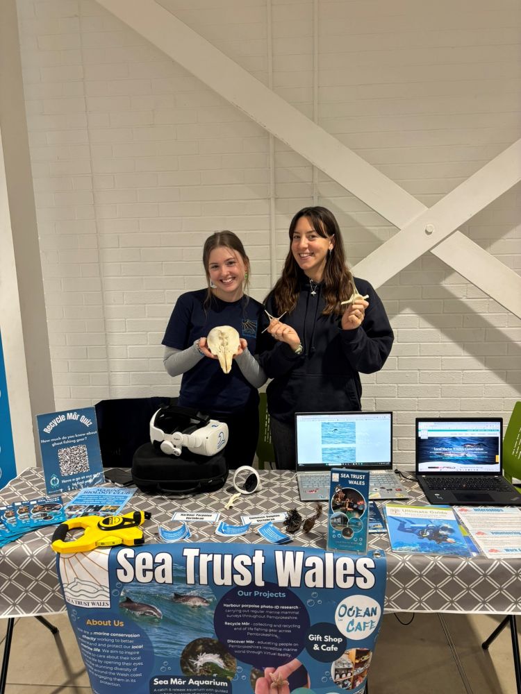 Two people are standing behind a table with exhibition material from Sea Trust Wales.