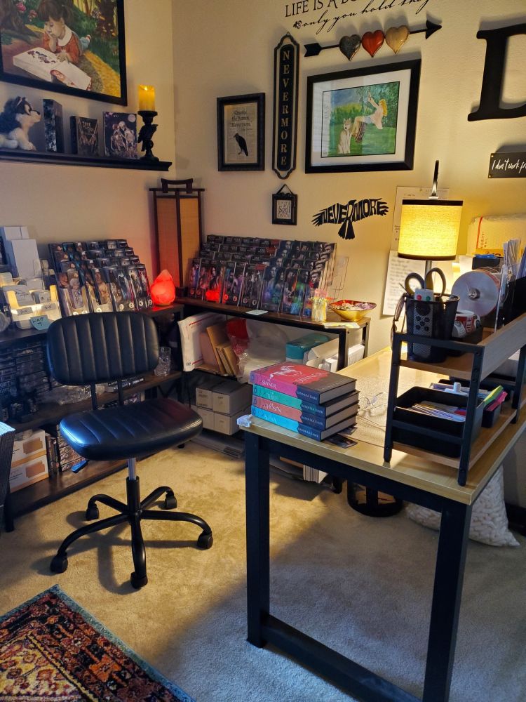 Office corner with narrow wall shelves filled with bookmarks, shipping supplies, and a salt lamp. The wall is covered with art, mostly raven and edgar allen poe-themed.