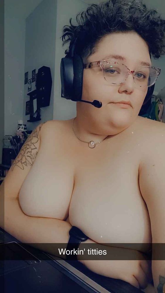 Thistle topless with her arm across her tits, Snapchat text overlay says "Workin' titties." She is wearing glasses and a headset with microphone.