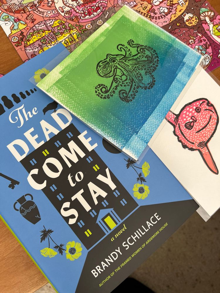 The cover of a book reads The Dead Come to Stay. It is a medium blue with neon green flowers and building accents. The two Lino prints are 1) a black octopus on blue and green overlapping rectangles and 2) a Mola mola colored in pink and orange gradient.