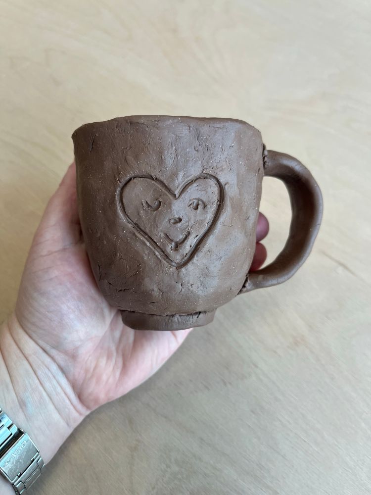 my light skinned hand holds a freshly made brown clay mug decorated with a heart shaped smiley face with a winking eye and tongue sticking out. background is a beige wooden table. 