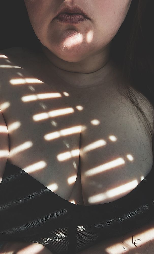A brunette goddess standing in the light coming through the blinds. The shadows of the light play over her breasts. She is wearing a black corset top that accentuates her full breasts. You can only see from her full lips down. Her hair spilling over her shoulder. 