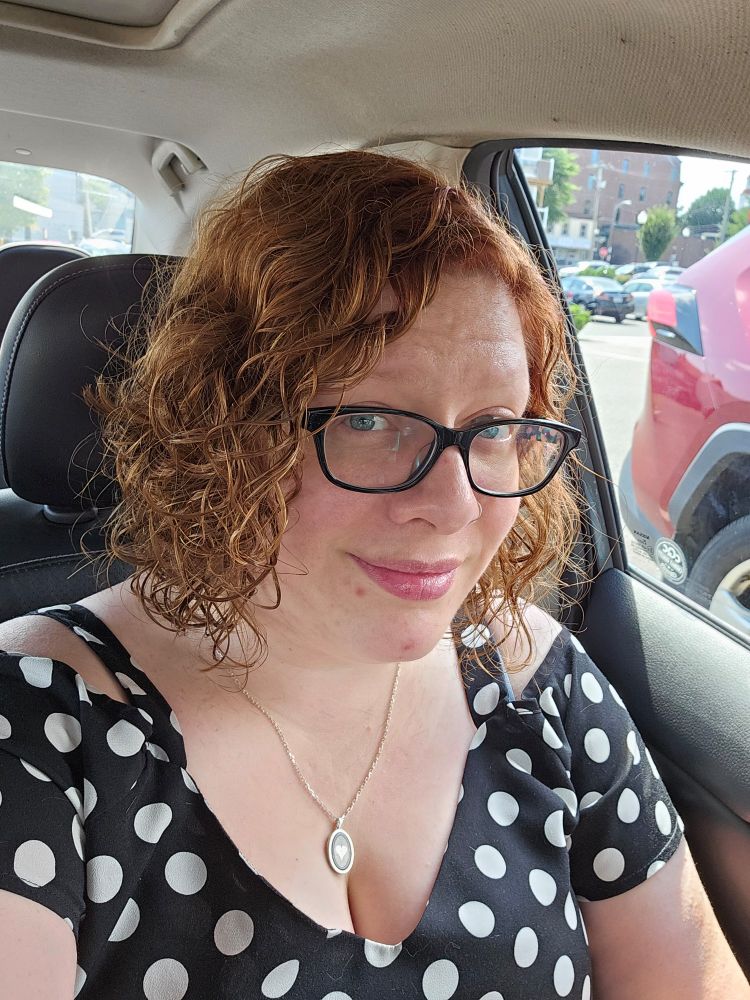 A red head, curly haired gal with glasses wearing a polka dot top