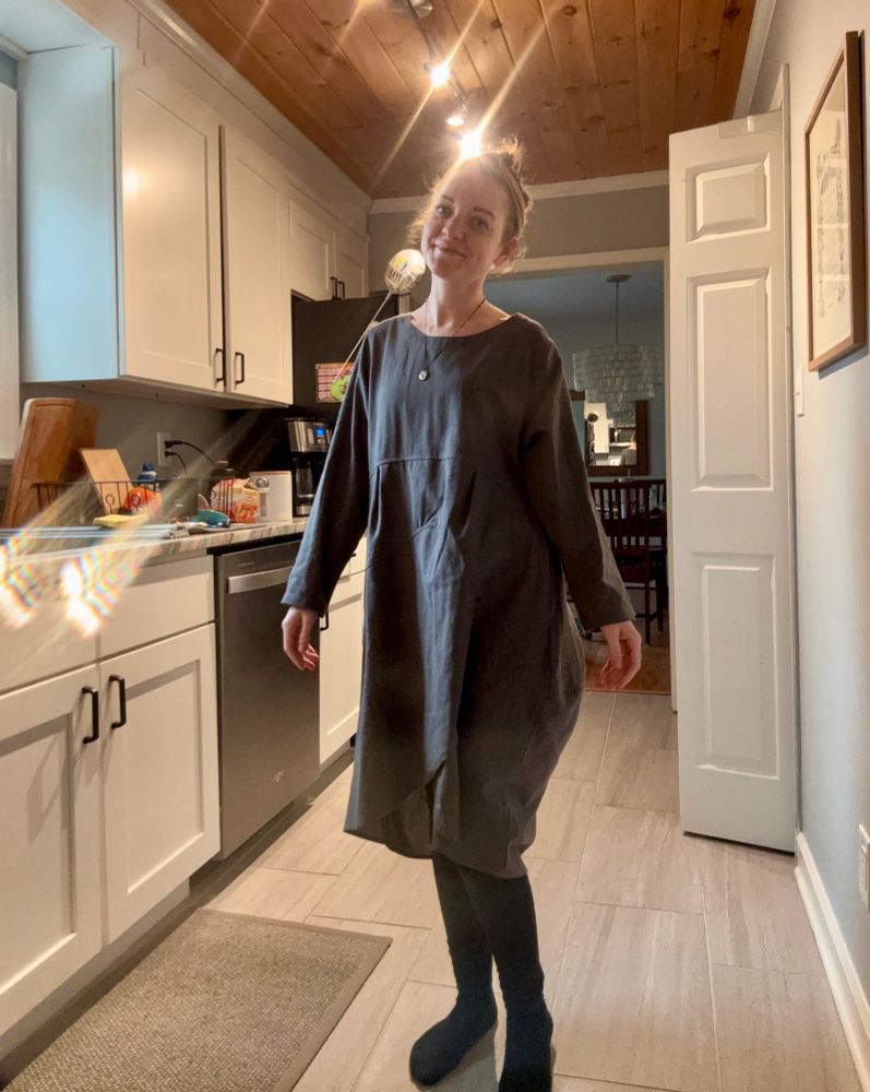 Em stands in a white and gray kitchen. A yellow lens flare is beside her. She is in a shapeless gray dress